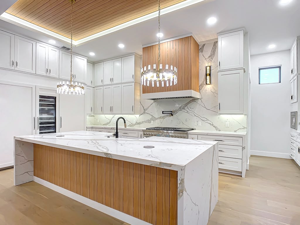 Finished kitchen with white cabinetry, marble waterfall island and wood ceiling accents