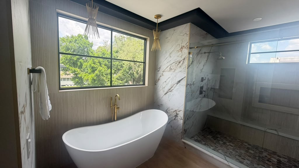 White freestanding tub beneath a black-framed window with gold pendant lights and glass shower